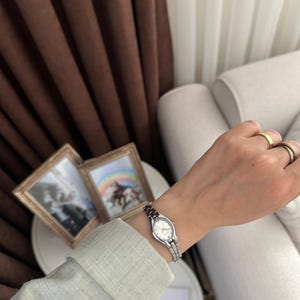Op de afbeelding: Een zilveren polshorloge met een witte wijzerplaat en een kleine, rechthoekige kast. Het horloge heeft een zilveren band met donkerrode accenten. Het horloge zit om een pols met gouden ringen. Twee ingelijste foto's staan op de achtergrond.