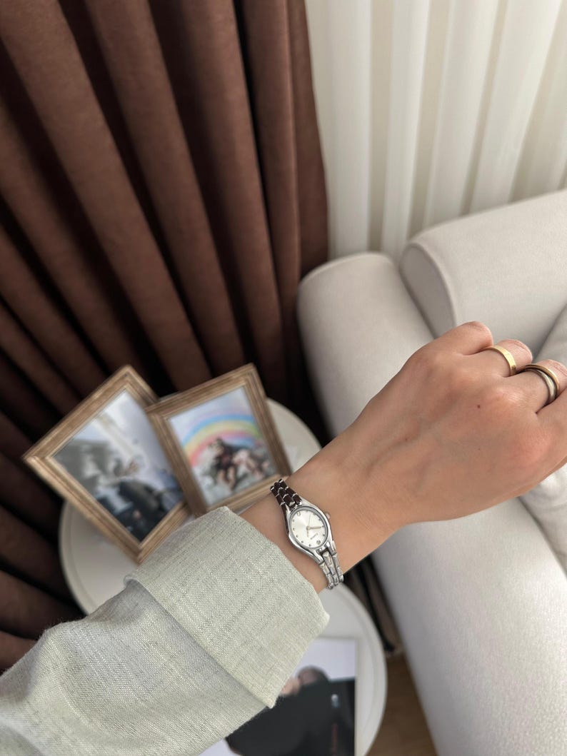 Op de afbeelding: Een zilveren polshorloge met een witte wijzerplaat en een kleine, rechthoekige kast. Het horloge heeft een zilveren band met donkerrode accenten. Het horloge zit om een pols met gouden ringen. Twee ingelijste foto's staan op de achtergrond.