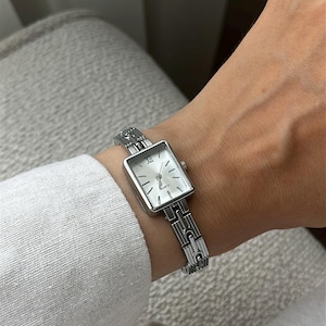 Puede incluir: Un reloj de pulsera plateado con una esfera cuadrada y una correa de eslabones a juego. La esfera del reloj es blanca con agujas plateadas y números romanos. El reloj se lleva en la muñeca.