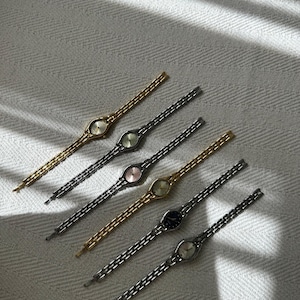 Op de afbeelding: Een assortiment delicate polshorloges in vintage-stijl. De horloges hebben ovale wijzerplaten en slanke schakelarmbanden in goud, zilver en zwart. De wijzerplaten tonen verschillende kleuren, waaronder goud, roze en zwart. De horloges zijn diagonaal gerangschikt op een getextureerd, gebroken witte ondergrond.