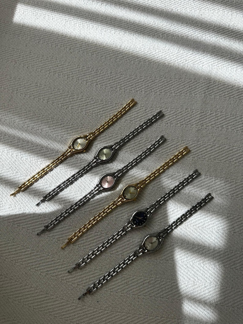 Op de afbeelding: Een assortiment delicate polshorloges in vintage-stijl. De horloges hebben ovale wijzerplaten en slanke schakelarmbanden in goud, zilver en zwart. De wijzerplaten tonen verschillende kleuren, waaronder goud, roze en zwart. De horloges zijn diagonaal gerangschikt op een getextureerd, gebroken witte ondergrond.
