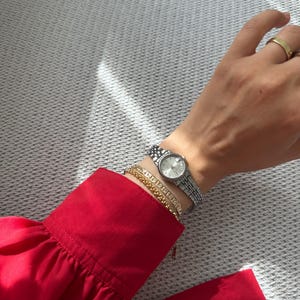 Può includere: Un orologio da polso argentato con quadrante rotondo e cinturino a maglie è indossato su un polso, insieme a due braccialetti d'oro. Il polso è accanto a una camicia rossa brillante a maniche lunghe. Un anello d'oro è su un dito.