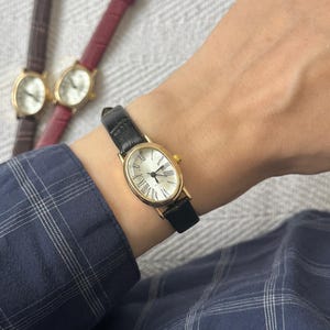 Könnte beinhalten: Eine goldfarbene Armbanduhr mit schwarzem Lederarmband wird am Handgelenk getragen. Die Uhr hat ein weißes ovales Zifferblatt mit römischen Ziffern. Zwei weitere Uhren mit braunen und weinroten Armbändern sind sichtbar.