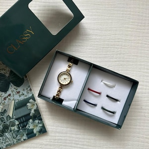 Puede incluir: Un reloj de pulsera dorado con esfera blanca y correa dorada a juego, presentado en una caja verde oscuro con la palabra "CLASSY" en letras doradas. La caja también contiene seis anillos de varios colores.
