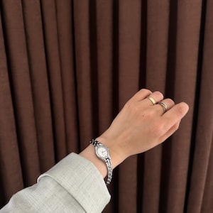 Op de afbeelding: Een zilveren polshorloge met een kleine, ovale witte wijzerplaat en een schakelband wordt om een pols gedragen. Het horloge wordt gecombineerd met twee ringen aan de vingers. De mouw van een lichtgroen overhemd is zichtbaar.