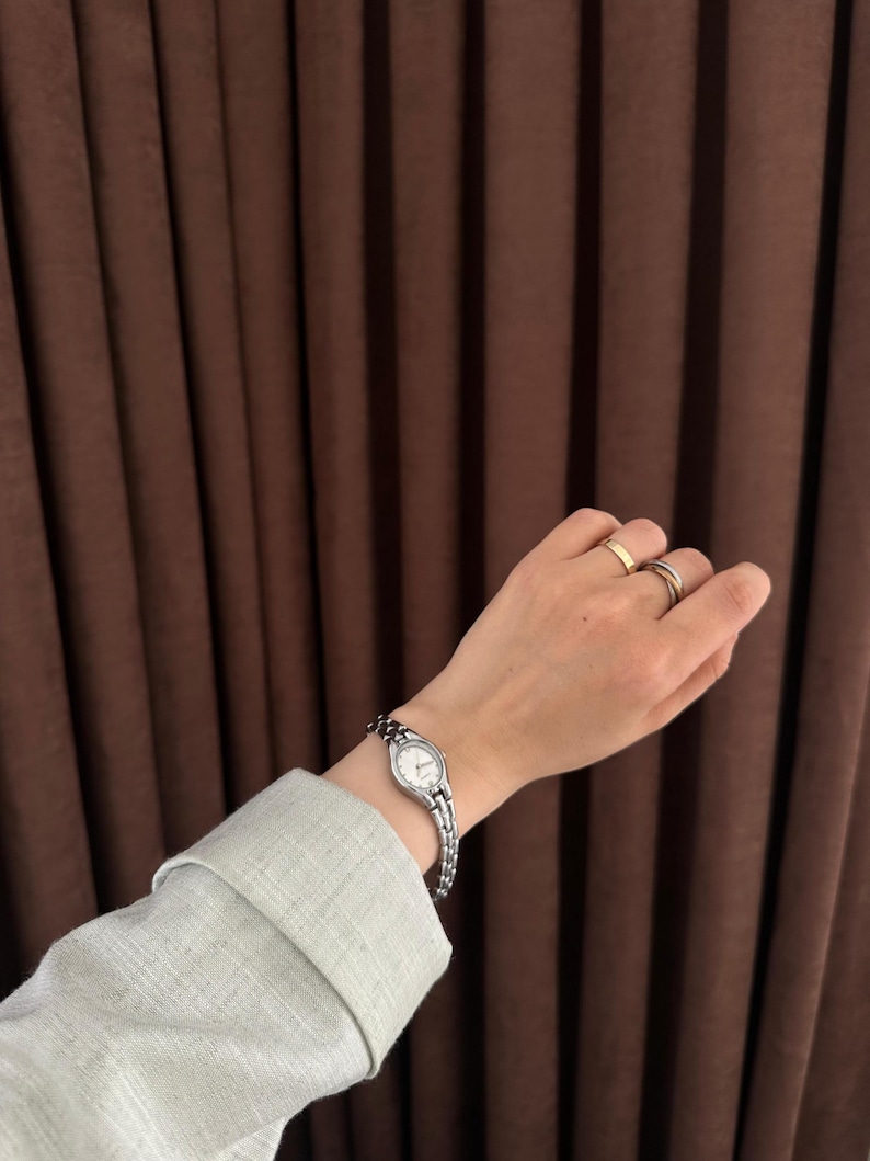 Op de afbeelding: Een zilveren polshorloge met een kleine, ovale witte wijzerplaat en een schakelband wordt om een pols gedragen. Het horloge wordt gecombineerd met twee ringen aan de vingers. De mouw van een lichtgroen overhemd is zichtbaar.