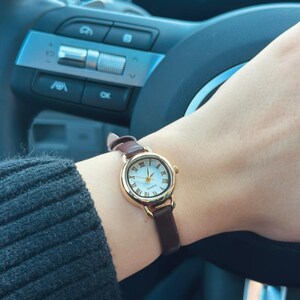 May include: A gold-toned wristwatch with a white face and Roman numeral hour markers. The watch has a brown leather band and is being worn on a wrist. The watch is a quartz movement.