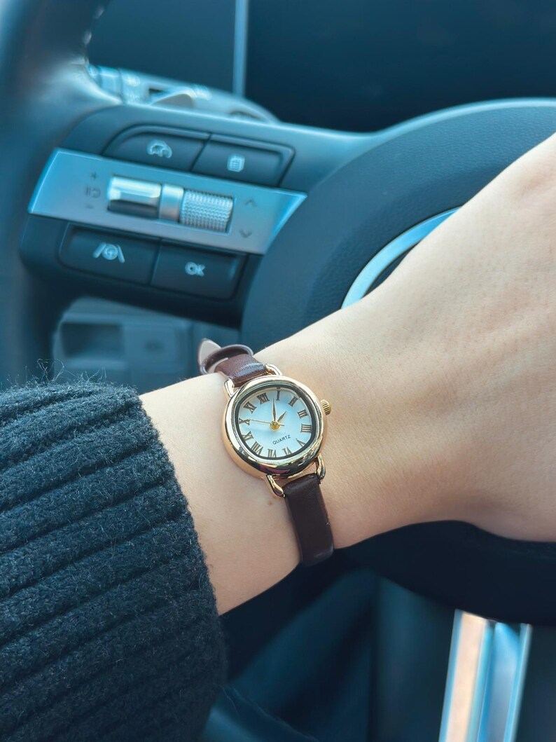 May include: A gold-toned wristwatch with a white face and Roman numeral hour markers. The watch has a brown leather band and is being worn on a wrist. The watch is a quartz movement.