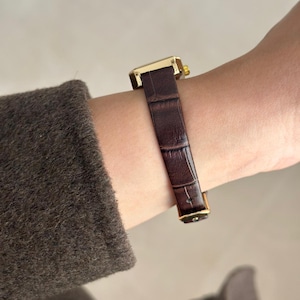 May include: A gold-toned rectangular wristwatch with a dark brown, textured leather band. The watch is fastened on a wrist, showcasing the band's intricate pattern and the watch's elegant design. The watch face is not visible.