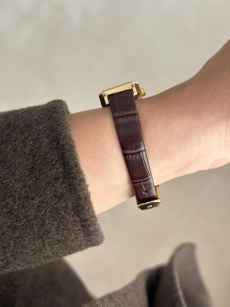 May include: A gold-toned rectangular wristwatch with a dark brown, textured leather band. The watch is fastened on a wrist, showcasing the band's intricate pattern and the watch's elegant design. The watch face is not visible.