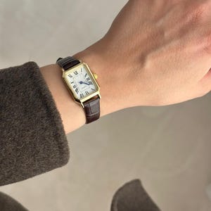 May include: A gold-toned rectangular wristwatch with a white face and Roman numeral hour markers. The watch has a brown leather band. The watch is approximately 1 inch wide and 1.5 inches long.