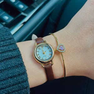 May include: A gold-toned watch with a brown leather band and a light blue face. A gold bracelet with a pink heart-shaped charm is also visible. The watch has Roman numerals and gold hands.