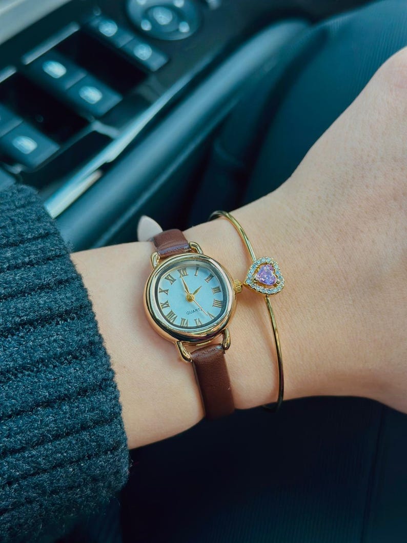May include: A gold-toned watch with a brown leather band and a light blue face. A gold bracelet with a pink heart-shaped charm is also visible. The watch has Roman numerals and gold hands.