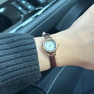 May include: A gold-toned wristwatch with a white face and Roman numeral hour markers. The watch has a brown leather band. The watch is on a wrist, with a gray sweater sleeve visible in the background.