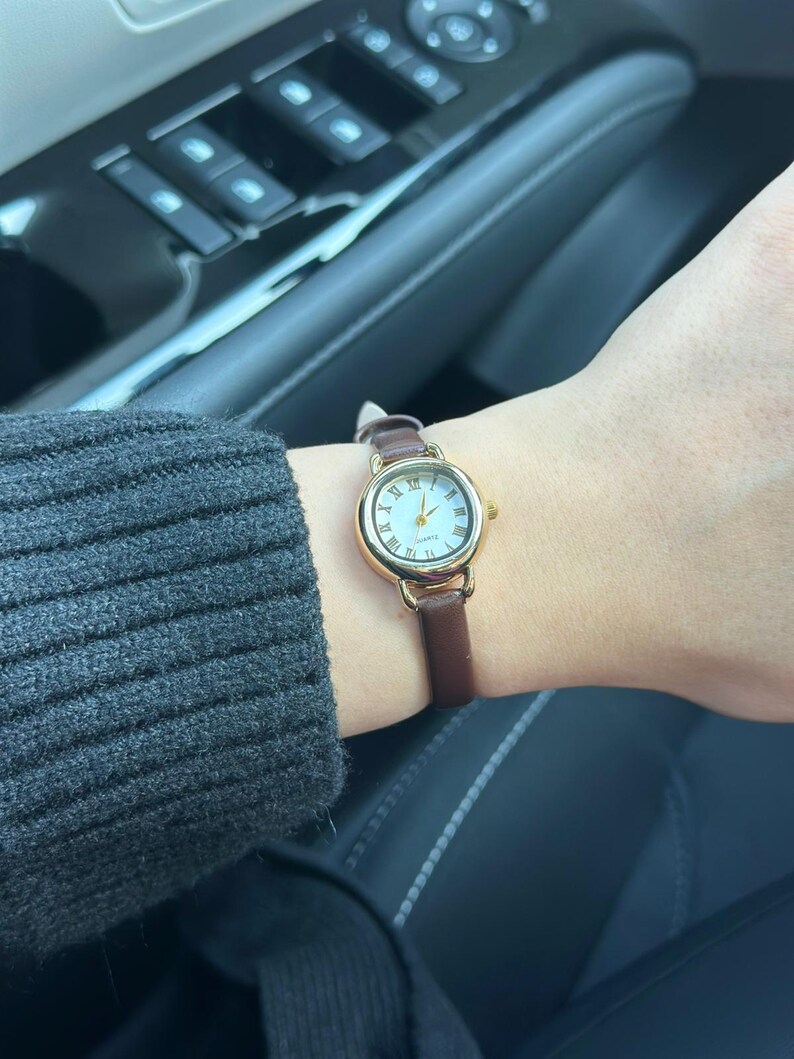 May include: A gold-toned wristwatch with a white face and Roman numeral hour markers. The watch has a brown leather band. The watch is on a wrist, with a gray sweater sleeve visible in the background.