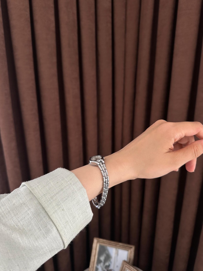 Op de afbeelding: Een zilverkleurig polshorloge met een rechthoekige wijzerplaat en een schakelband wordt om een pols gedragen. Het horloge wordt getoond tegen een achtergrond van een lichtgroene mouw en een bruin gordijn.
