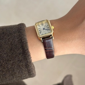 May include: A gold-toned square wristwatch with a white face and Roman numeral hour markers. The watch has a dark brown leather band. The watch is on a wrist.