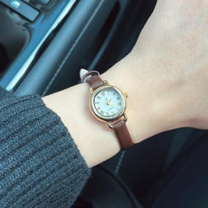 May include: A gold-toned wristwatch with a white face and Roman numeral hour markers. The watch has a brown leather band. The watch is on a wrist, with a gray sweater sleeve visible.