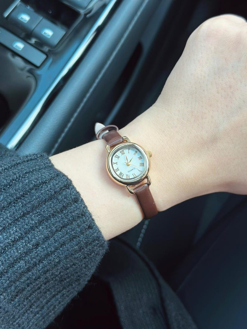 May include: A gold-toned wristwatch with a white face and Roman numeral hour markers. The watch has a brown leather band. The watch is on a wrist, with a gray sweater sleeve visible.