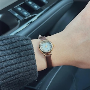 May include: A gold-toned wristwatch with a white face and Roman numeral hour markers. The watch has a brown leather band. The watch is on a wrist, with a dark gray sweater sleeve visible in the background.