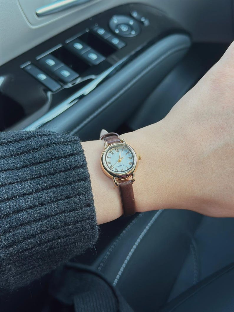 May include: A gold-toned wristwatch with a white face and Roman numeral hour markers. The watch has a brown leather band. The watch is on a wrist, with a dark gray sweater sleeve visible in the background.