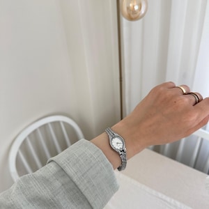Op de afbeelding: Een zilveren polshorloge met een witte wijzerplaat wordt om een pols gedragen. Het horloge heeft een schakelband en een kleine, ronde wijzerplaat. De persoon draagt een grijs shirt met lange mouwen. De achtergrond omvat een armatuur en een witte stoel.