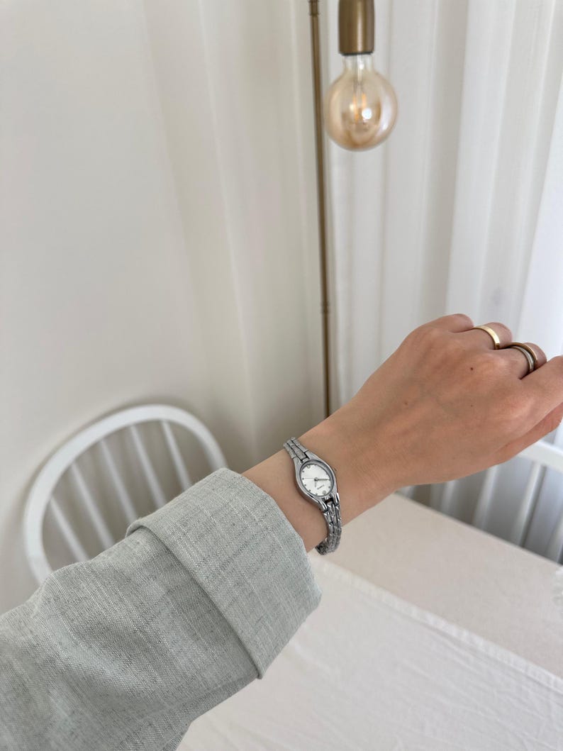 Op de afbeelding: Een zilveren polshorloge met een witte wijzerplaat wordt om een pols gedragen. Het horloge heeft een schakelband en een kleine, ronde wijzerplaat. De persoon draagt een grijs shirt met lange mouwen. De achtergrond omvat een armatuur en een witte stoel.