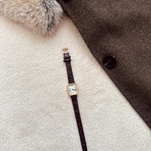 May include: A rectangular gold-toned wristwatch with a dark brown leather band lies on a white textured surface. The watch face is white with black Roman numerals and blue hands. A dark brown coat with black buttons and a fur collar are in the background.