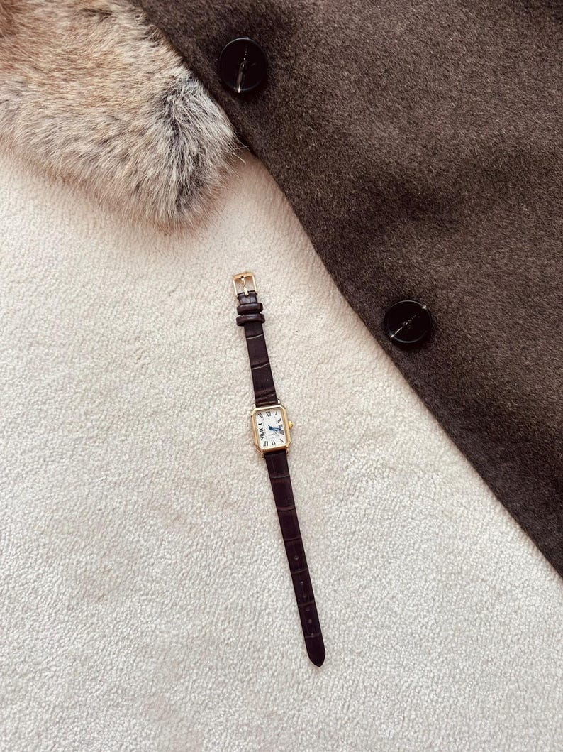 May include: A rectangular gold-toned wristwatch with a dark brown leather band lies on a white textured surface. The watch face is white with black Roman numerals and blue hands. A dark brown coat with black buttons and a fur collar are in the background.