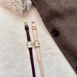 May include: Two rectangular gold-toned watches with leather straps. One watch has a dark brown strap, the other a light pink strap. The watch faces have Roman numerals and hands. The watches are on a white textured surface.