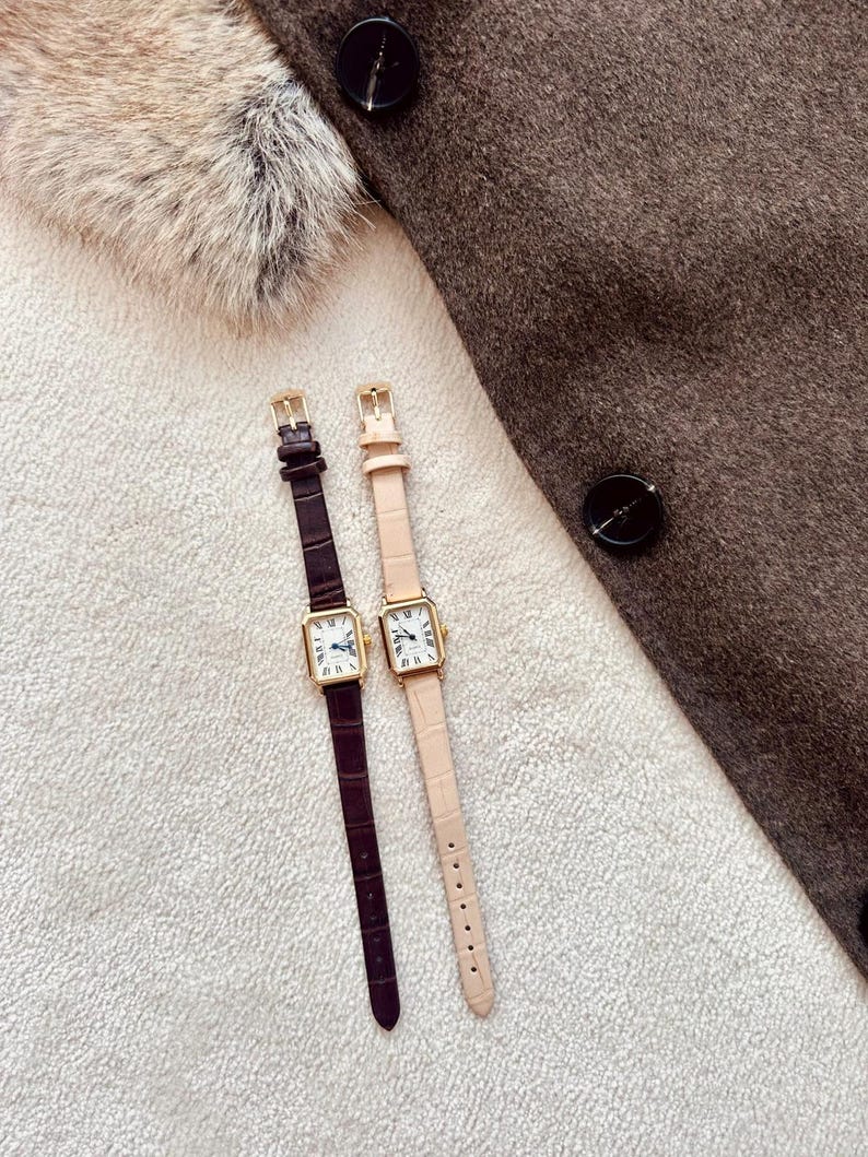 May include: Two rectangular gold-toned watches with leather straps. One watch has a dark brown strap, the other a light pink strap. The watch faces have Roman numerals and hands. The watches are on a white textured surface.