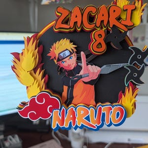 May include: A colorful cake topper featuring the character Naruto from the anime series. The topper includes the name "Zacari" and the number "8" in orange, with flames and ninja stars. The word "Naruto" is also displayed.