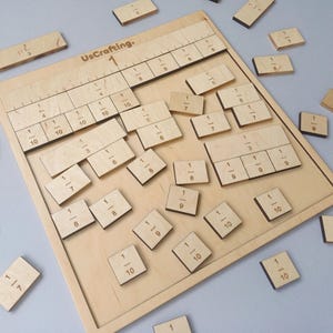 Può includere: Una tavola di frazioni in legno con vari pezzi frazionari. La tavola presenta linee e numeri incisi, comprese frazioni come 1/2, 1/3 e 1/4. Il testo "UsCrafting" è visibile. I pezzi sono sparsi, suggerendo un'attività di apprendimento.