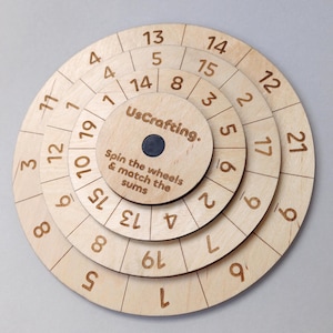 May include: A wooden educational toy with concentric rotating wheels, each marked with numbers. The center wheel has the text "UsCrafting." and "Spin the wheels & match the sums." The numbers are etched into the light-coloured wood.