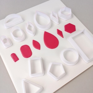 May include: An assortment of white plastic cookie cutters in various shapes, including circles, teardrops, and geometric forms, alongside red clay cutouts. The cutters are arranged on a white surface, providing a contrast to the vibrant red clay.