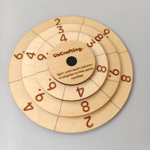 May include: A wooden multiplication wheel with concentric circles and numbers. The center circle has the text "UsCrafting." and instructions. The numbers are in a brown font. The wheel is designed for educational purposes.