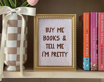 Peut inclure: Une étagère en bois présente des livres colorés, un vase à carreaux beige et blanc avec des tulipes roses, et une pancarte dorée avec l'inscription "BUY ME BOOKS & TELL ME I'M PRETTY".
