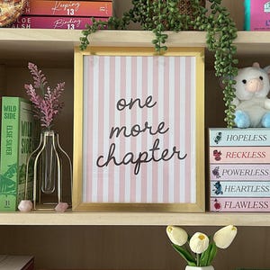 May include: A bookshelf displays various books, decorative items, and a framed print. The print reads "one more chapter." Books with colorful spines and titles like "Betting on You" and "Legacy" are visible. A decorative orange juice carton vase is also present.