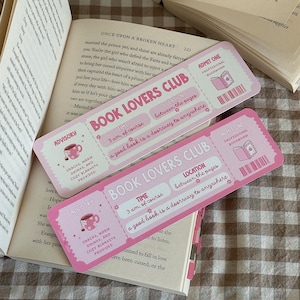 May include: Two pink Book Lovers Club ticket-style bookmarks with text, resting on an open book. The bookmarks have text that reads "3 am, of course" and "a good book is a doorway to anywhere."