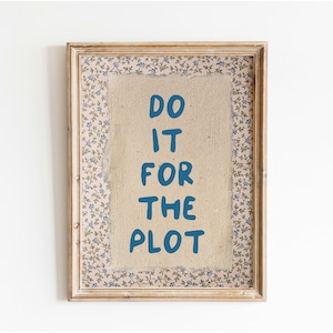 May include: A framed print with a light brown wooden frame and a white background. The print features the text "DO IT FOR THE PLOT" in blue lettering.