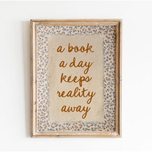 May include: A framed print with a floral border and the text "a book a day keeps reality away" in orange lettering on a beige background.