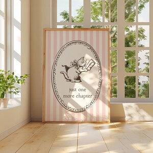 May include: A framed print with a cartoon cat reading a book, set against a pink and white striped background. The brown cat illustration is within an oval frame, with the text "just one more chapter" below. The frame is wooden.