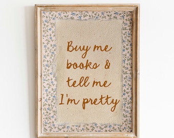 May include: A framed wall art piece with the text "Buy me books & tell me I'm pretty" in brown script. The artwork features a beige canvas center, a floral patterned border with blue flowers, and a distressed wooden frame.