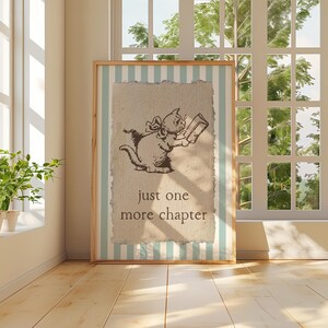 May include: A framed print featuring a cat reading a book, with the text "just one more chapter." The artwork has a light brown background with a light blue and white striped border. The frame is made of wood.