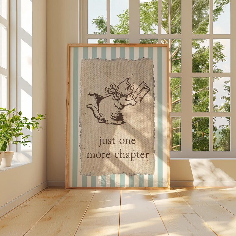 May include: A framed print featuring a cat reading a book, with the text "just one more chapter." The artwork has a light brown background with a light blue and white striped border. The frame is made of wood.