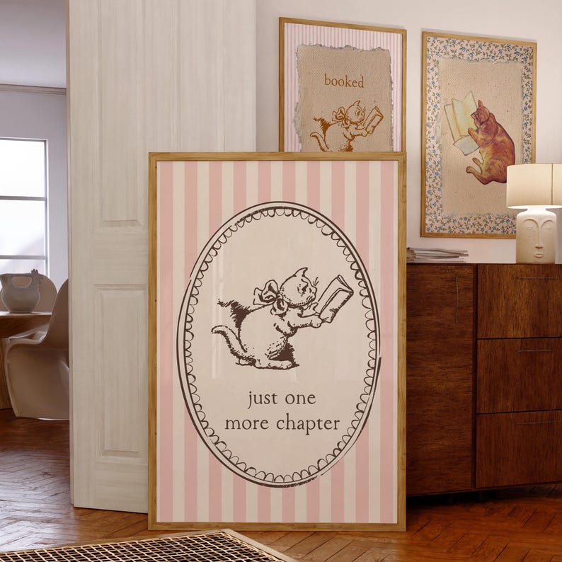 May include: A framed print with a pink and white striped background features a cartoon cat reading a book, with the text "just one more chapter." Two other framed prints with cat illustrations are visible.