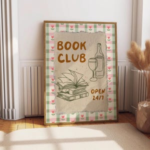 May include: A brown framed print with a white background and green and pink gingham border. The print features a hand-drawn illustration of a stack of books, a glass of wine, and a bottle of wine. The text "BOOK CLUB OPEN 24/7" is written in brown on the print.