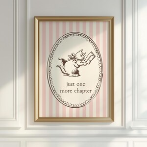 May include: A framed print with a gold-coloured frame, featuring a cartoon cat reading a book. The text "just one more chapter" is below the cat. The background has pink and white vertical stripes. The cat and text are brown.
