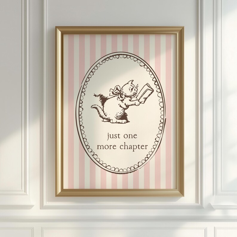 May include: A framed print with a gold-coloured frame, featuring a cartoon cat reading a book. The text "just one more chapter" is below the cat. The background has pink and white vertical stripes. The cat and text are brown.