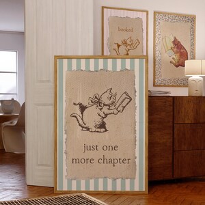 May include: Three framed art prints featuring illustrations of cats reading books. The largest print has a brown cat illustration with the text "just one more chapter" on a canvas-like background. The frame has a light blue and white striped border.